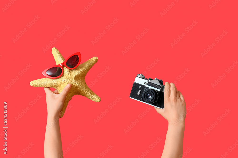 Female hands with camera and starfish in sunglasses on red background