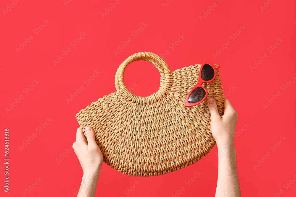 Male hands with wicker bag and sunglasses on red background