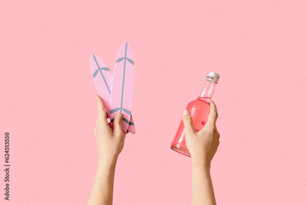 Female hands with mini surfboards and bottle of cocktail on pink background