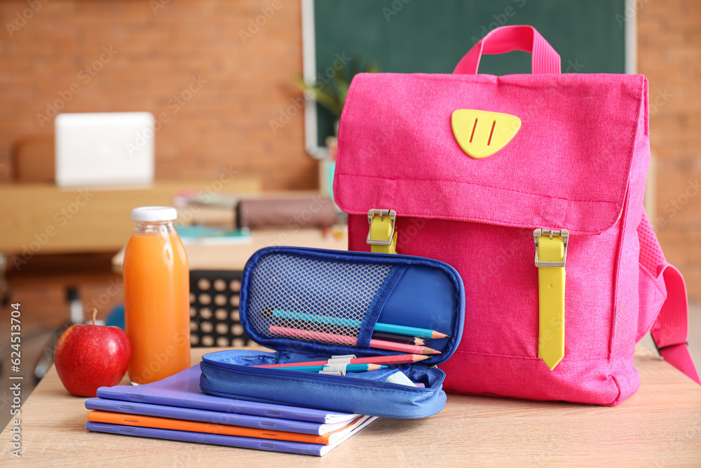 Backpack with bottle of juice, fresh apple and stationery on desk in classroom