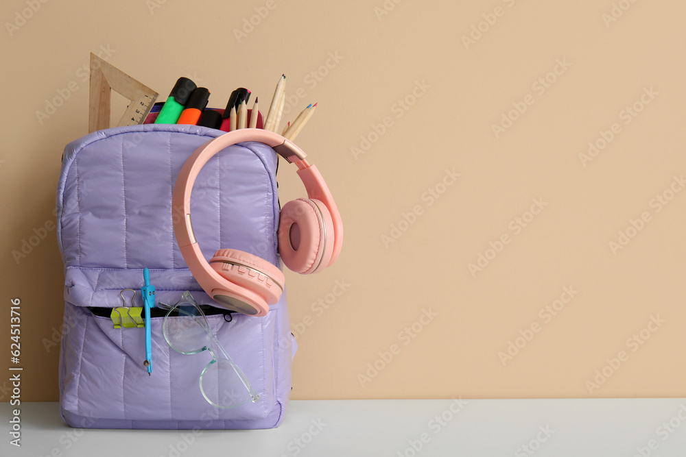 Lilac school backpack with different stationery and headphones on white table near orange wall