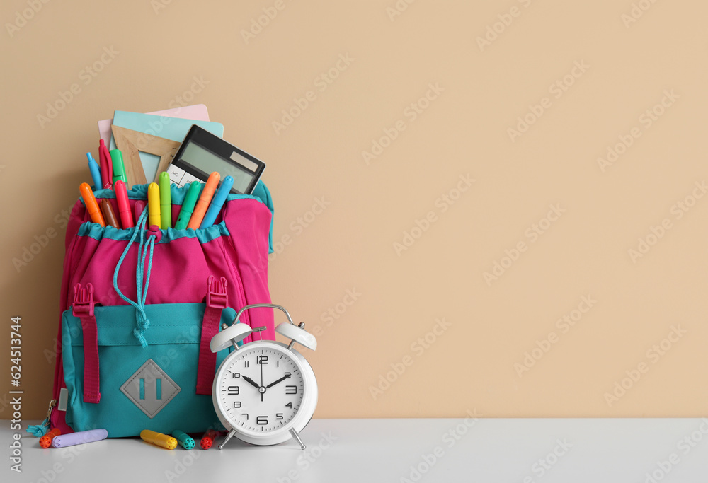 Colorful school backpack with different stationery and alarm clock on white table near orange wall