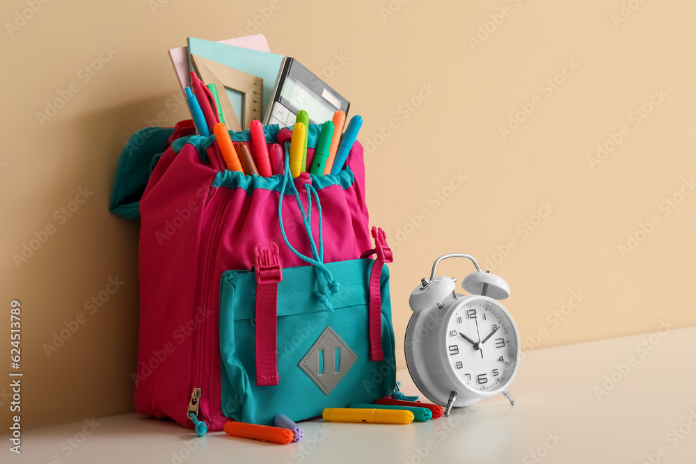 Colorful school backpack with different stationery and alarm clock on white table near orange wall