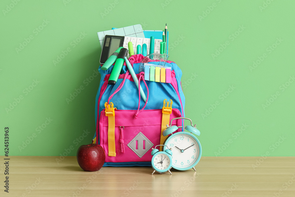 Colorful school backpack with different stationery and alarm clocks on wooden table near green wall