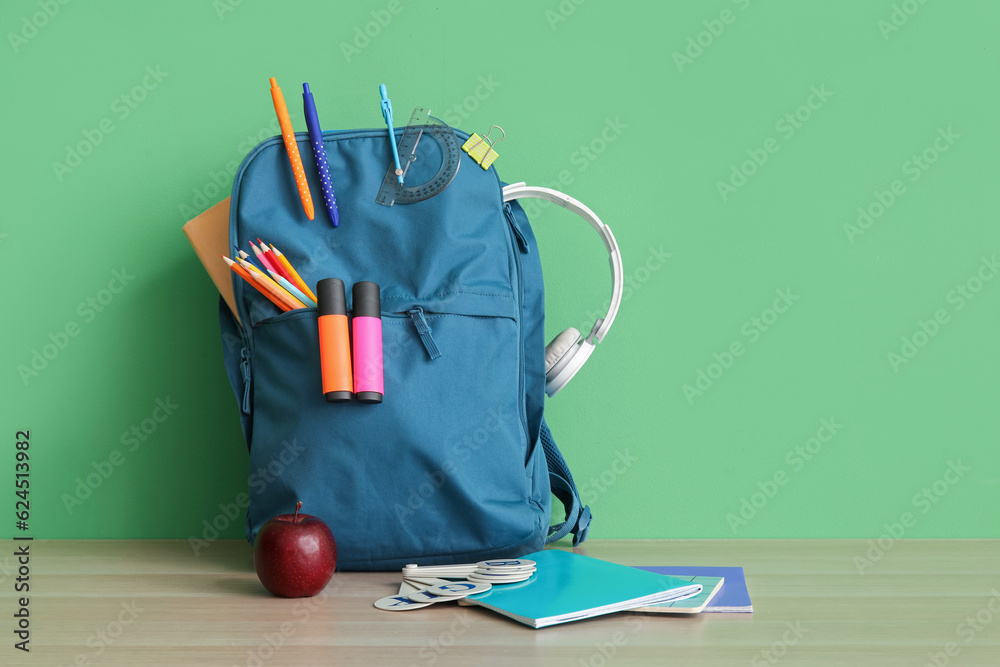 Blue school backpack with different stationery and headphones on wooden table near green wall