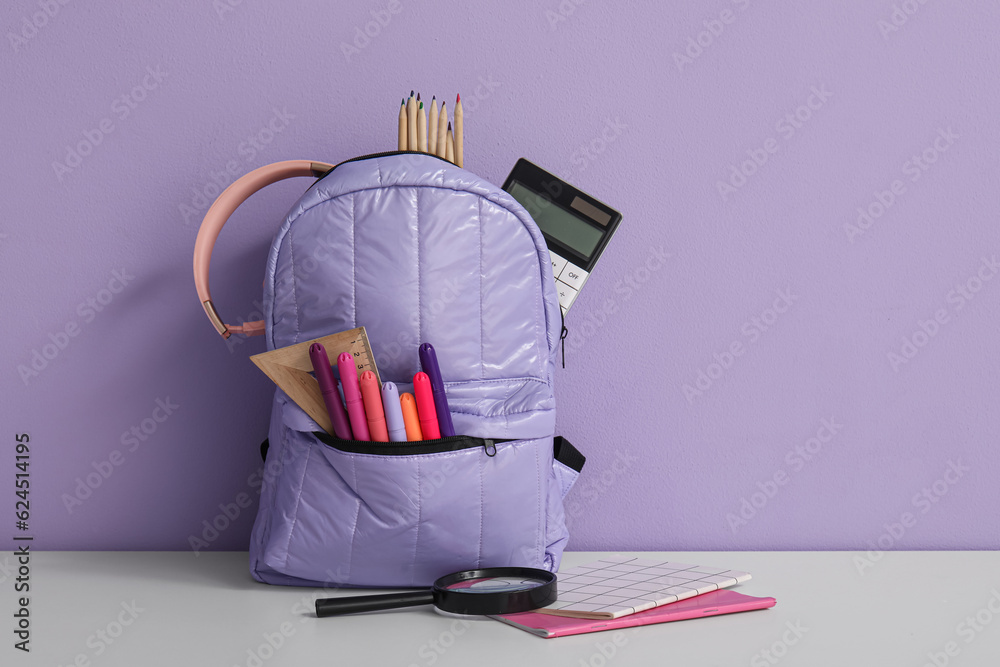 School backpack with different stationery and headphones on white table near purple wall