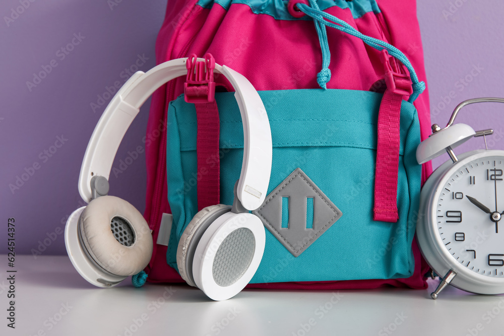 Colorful school backpack with headphones and alarm clock on white table near purple wall