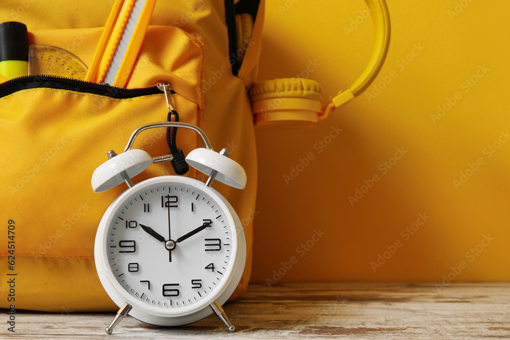 School backpack with stationery, headphones and alarm clock on wooden table near yellow wall