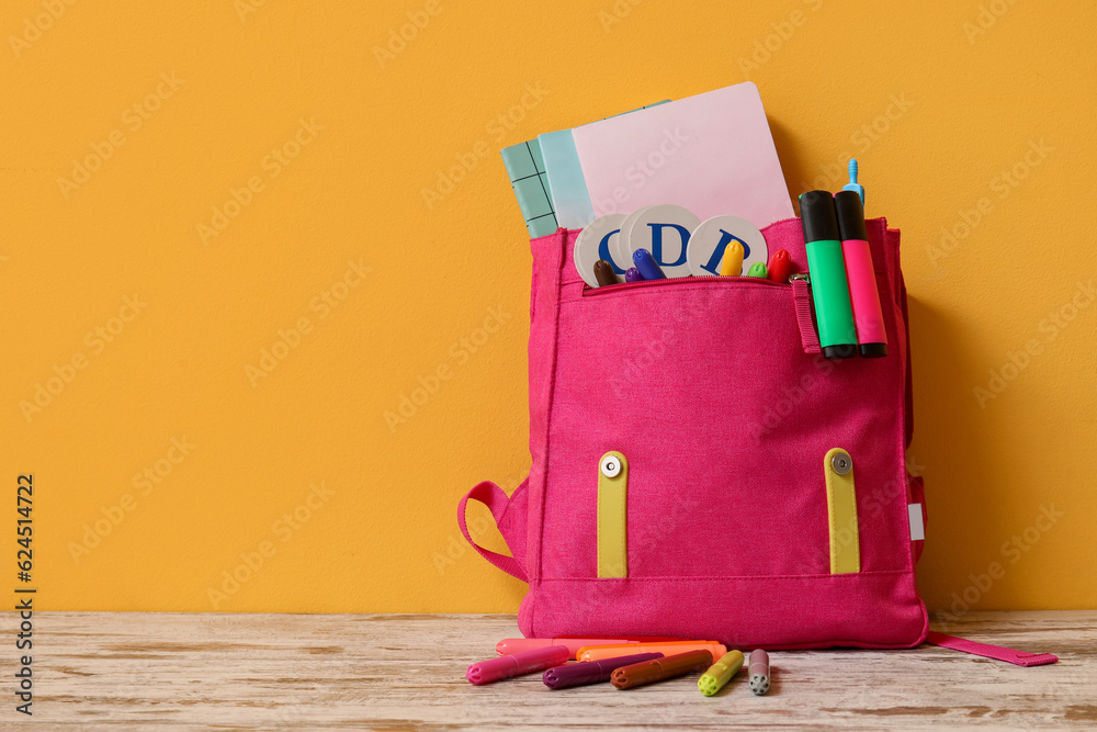 Pink backpack with different stationery on wooden table near yellow wall