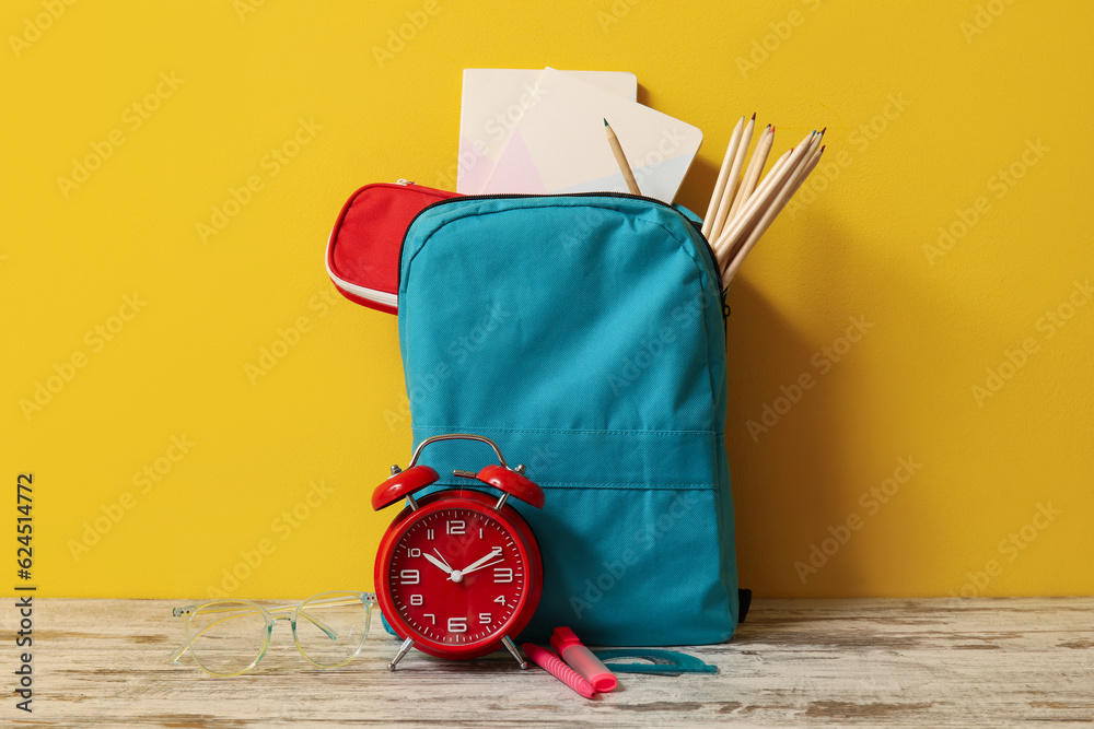 Blue school backpack with different stationery and alarm clock on wooden table near yellow wall