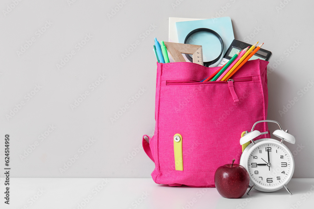 Pink school backpack with different stationery, alarm clock and fresh apple on white table near grey