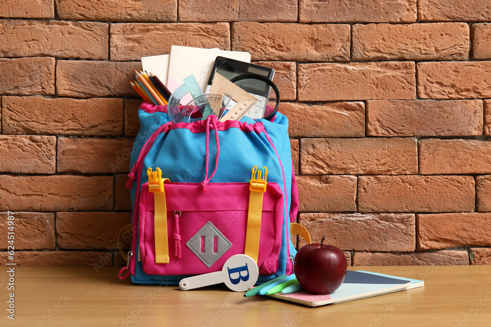 Colorful school backpack with different stationery on wooden table near brown brick wall