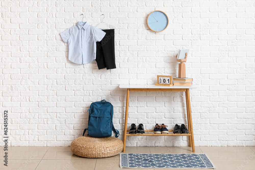 Table with shoes, backpack, clock and stylish school uniform hanging on light brick wall in room