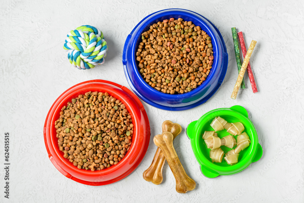 Bowls with dry dog food, treats and toy ball on light background