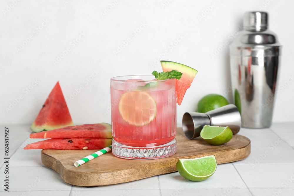 Board with glass of tasty watermelon cocktail, shaker, jigger and lime on white tile table