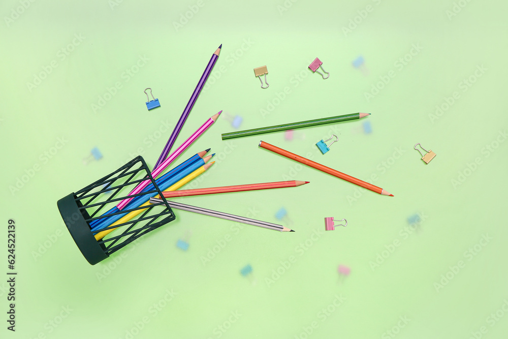 Overturned pen cup with colorful pencils and paperclips on green background