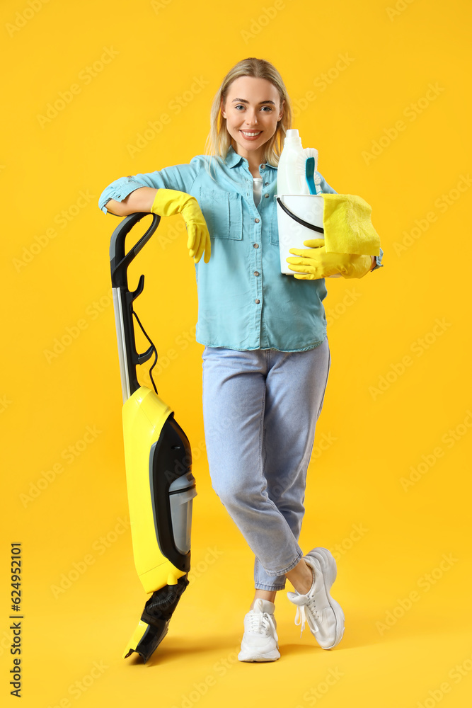 Young woman with hoover and cleaning supplies on yellow background