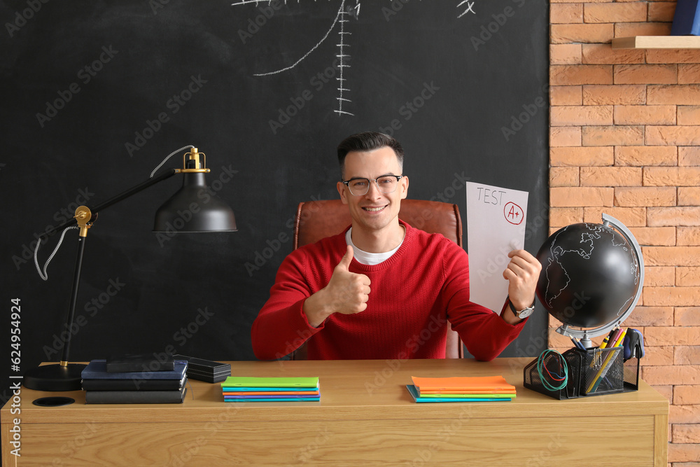 Male teacher with checked test sheet showing thumb-up at table in classroom