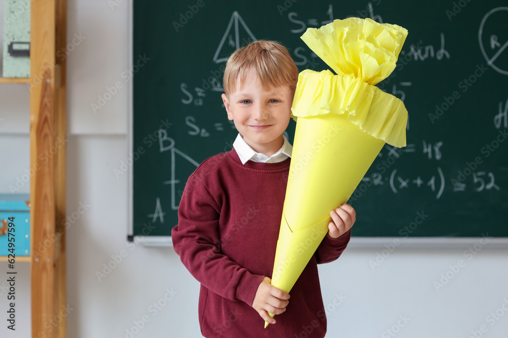 Happy little boy with yellow school cone in classroom near blackboard