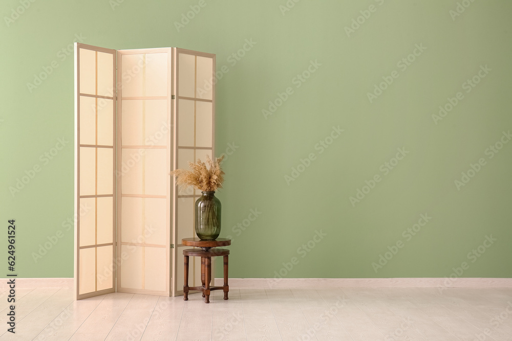 Folding screen, chair and vase of dried flowers near green wall