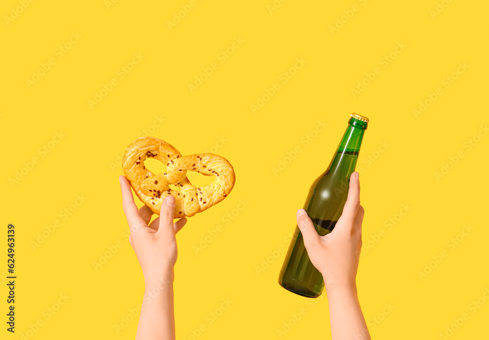 Female hands with bottle of cold beer and pretzel on yellow background