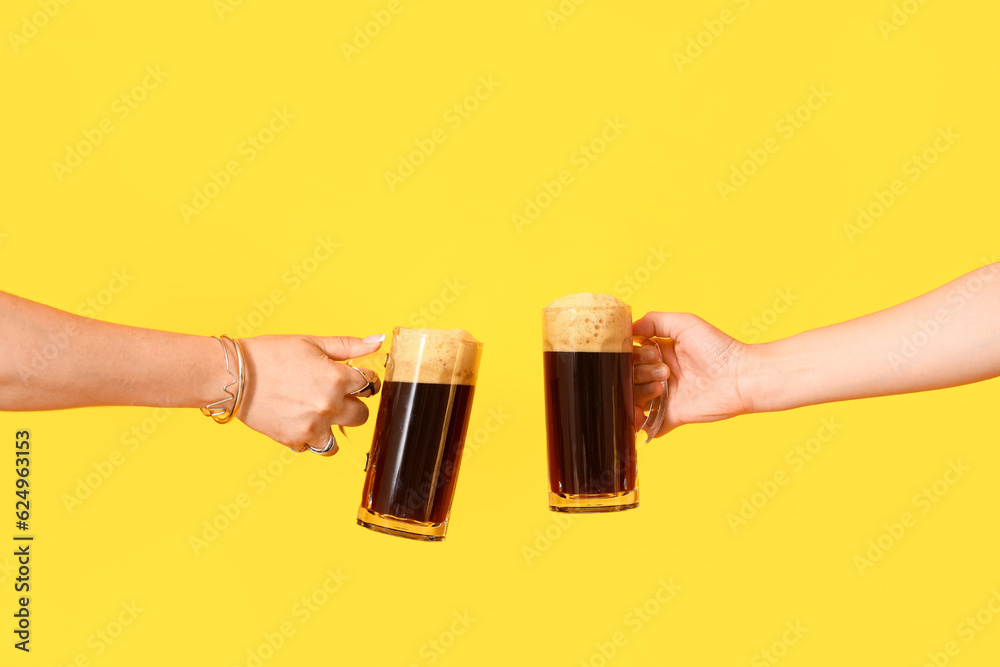 Female hands with mugs of cold beer on yellow background