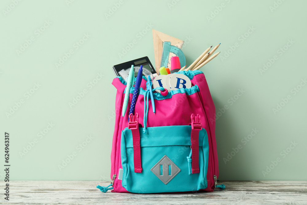 Colorful school backpack with different stationery on wooden table near pale green wall