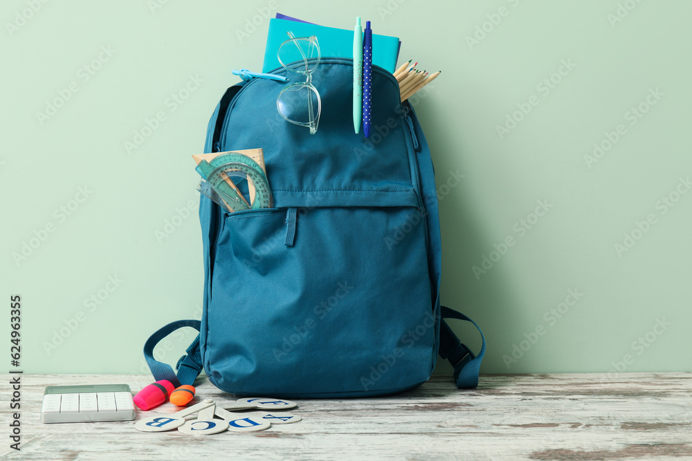 Blue school backpack with different stationery and calculator on wooden table near pale green wall