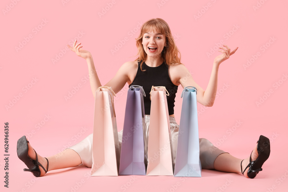 Beautiful happy young woman with shopping bags sitting on pink background