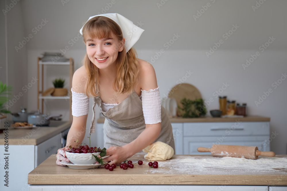 Beautiful young woman with bowl of cherries, rolling pin and dough on table in kitchen