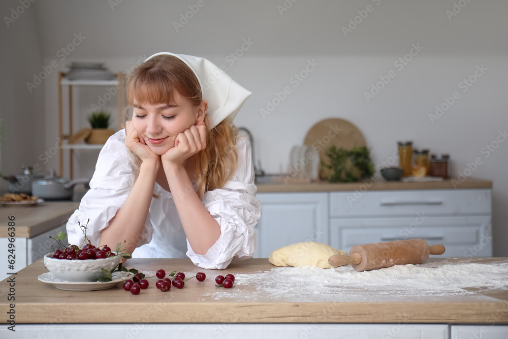 Beautiful young woman with bowl of cherries, rolling pin and dough on table in kitchen