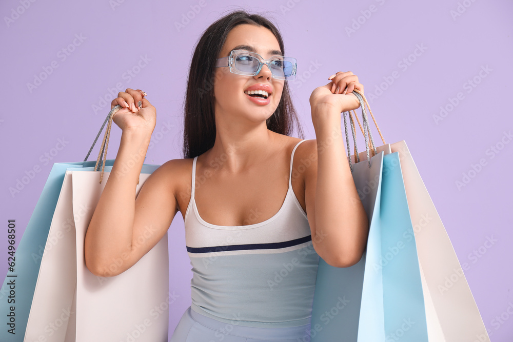 Happy beautiful young woman with shopping bags on lilac background