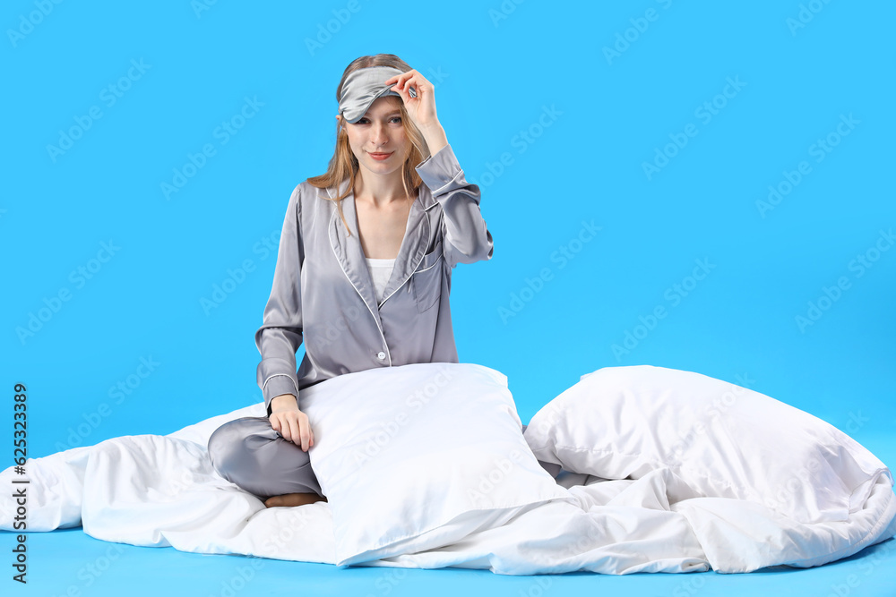 Young woman in pajamas with pillows and blanket on blue background