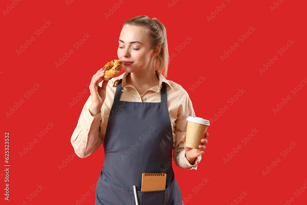 Young barista with cup of coffee eating bun on red background