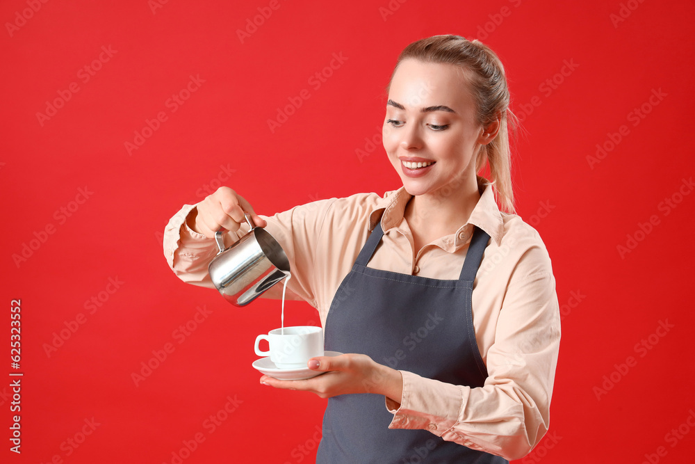 Young barista pouring milk into cup of coffee on red background