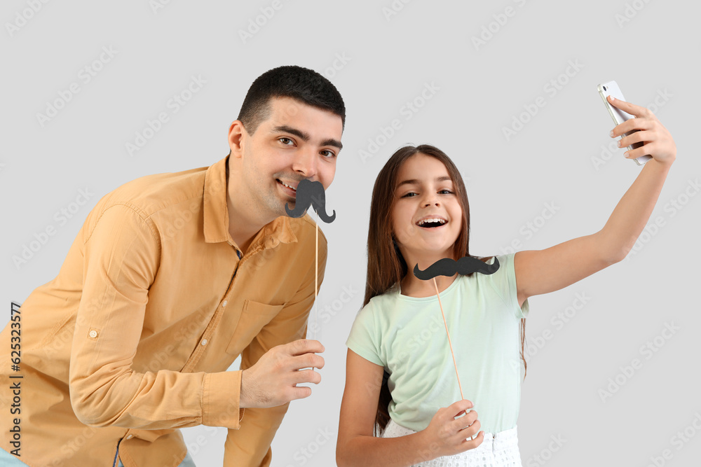 Portrait of father and his little daughter with paper mustache taking selfie on light background