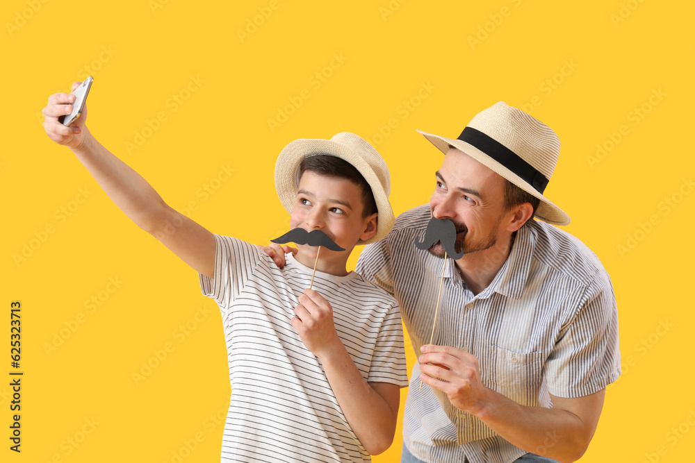 Portrait of father and his little son with paper mustache taking selfie on yellow background