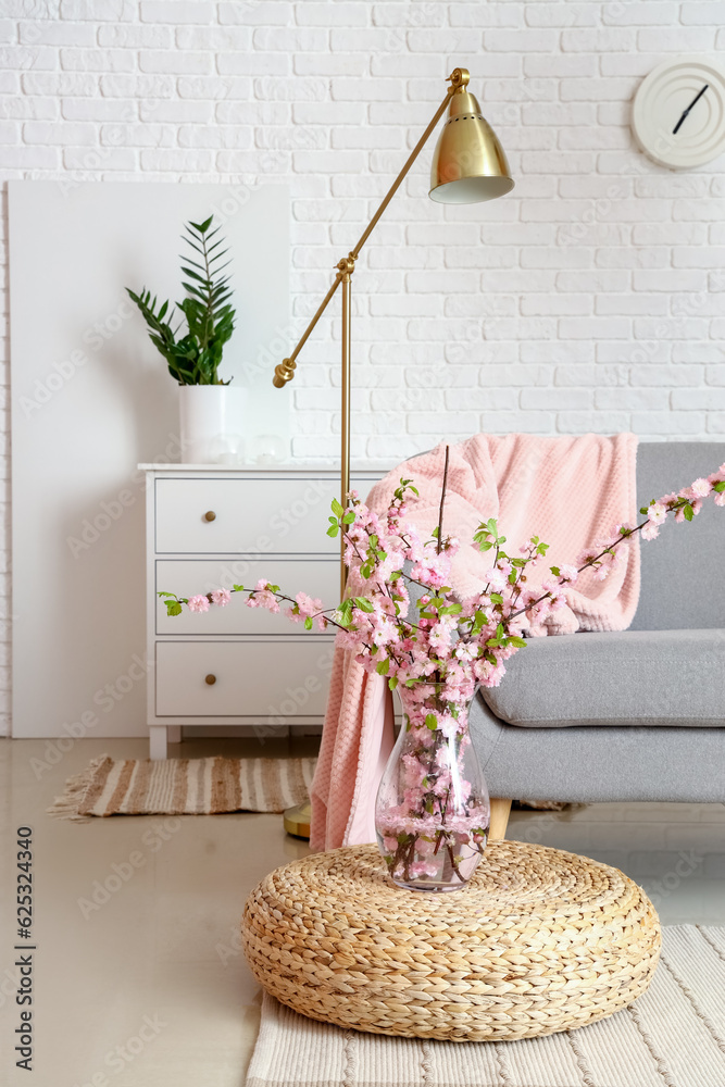 Vase with blooming sakura branches near grey sofa in interior of living room