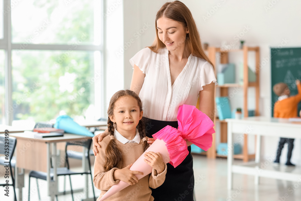 Happy little girl with pink school cone and teacher in classroom