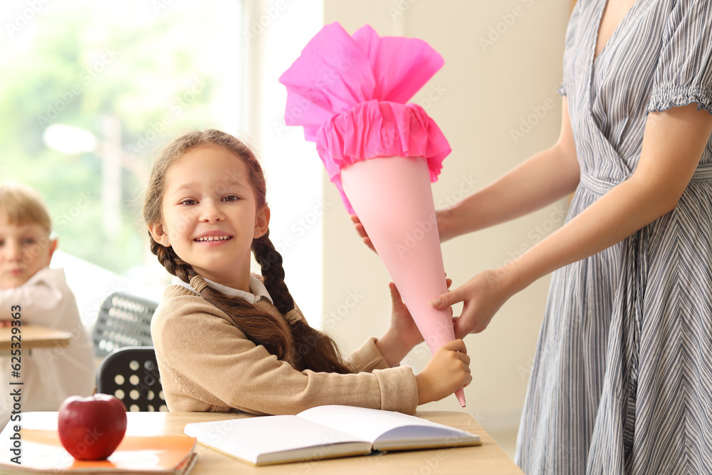 Teacher greeting her happy pupil with pink school cone in classroom