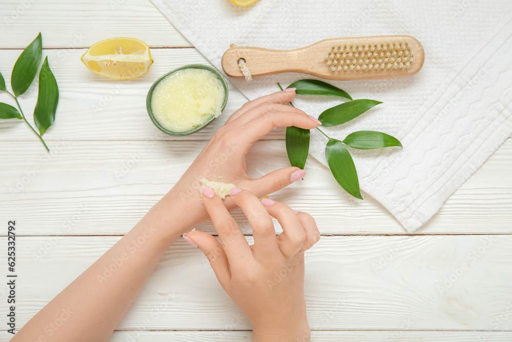 Woman with massage brush applying lemon body scrub on her body against white wooden background