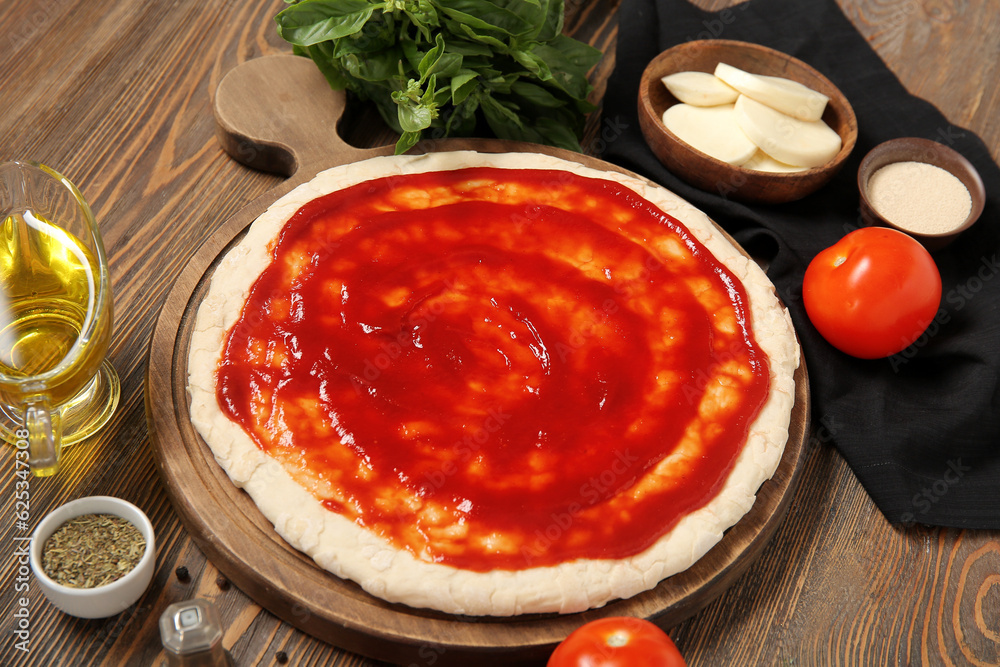 Board of pizza dough with tomato sauce and ingredients on wooden background