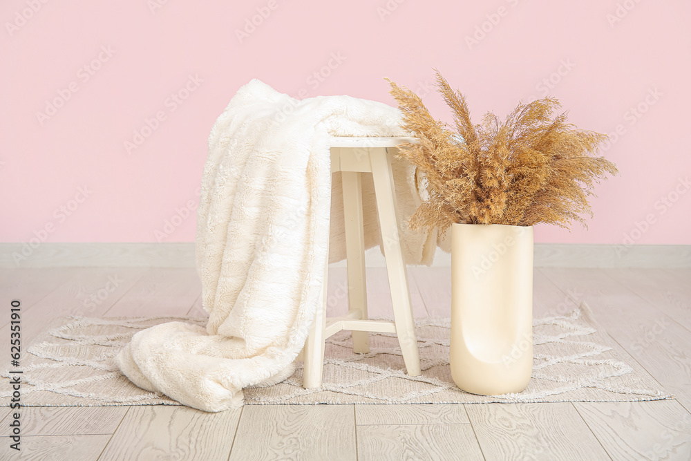 Blanket on stool and vase with pampas grass near pink wall