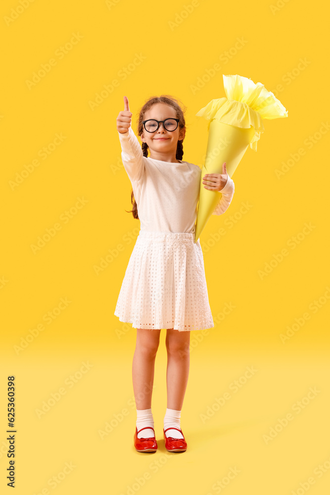 Happy little girl in eyeglasses with school cone showing thumb-up gesture on yellow background