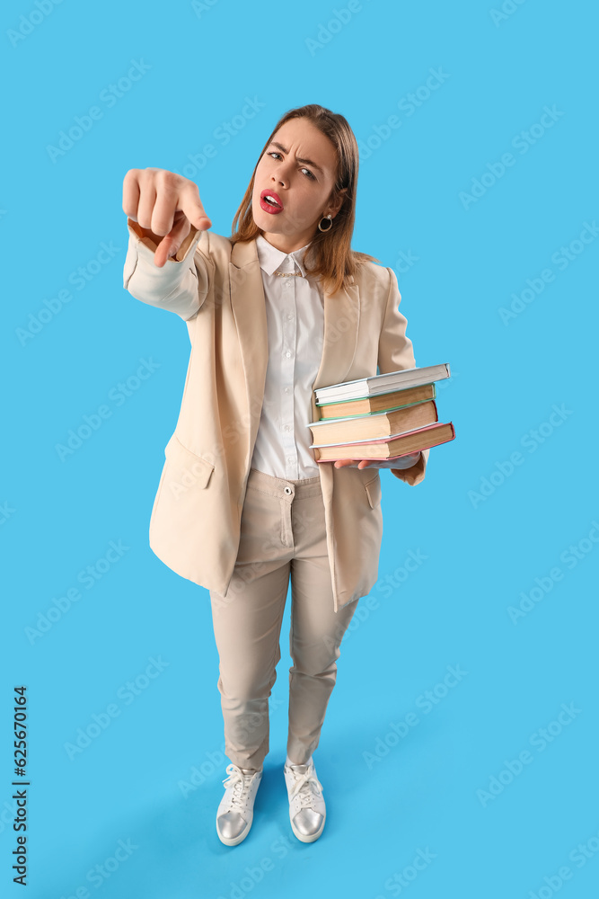 Upset young woman with books pointing at viewer on blue background