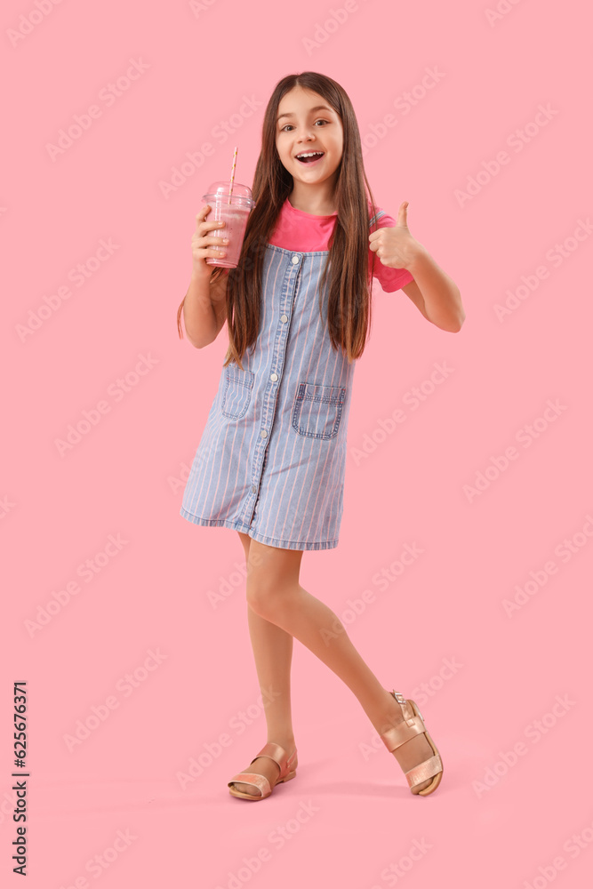 Little girl with glass of smoothie showing thumb-up on pink background