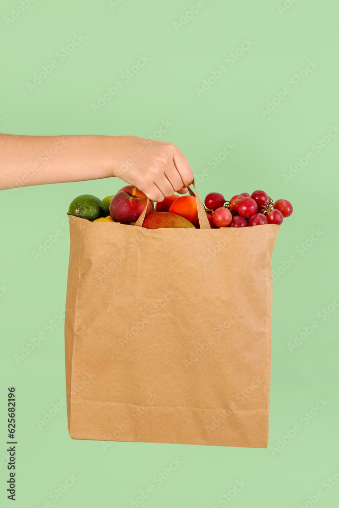 Female hand with paper bag full of fresh fruits on green background