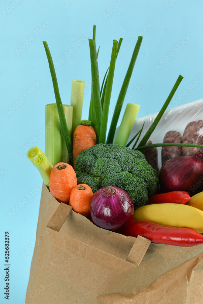 Paper bag full of fresh vegetables and sausages on blue background