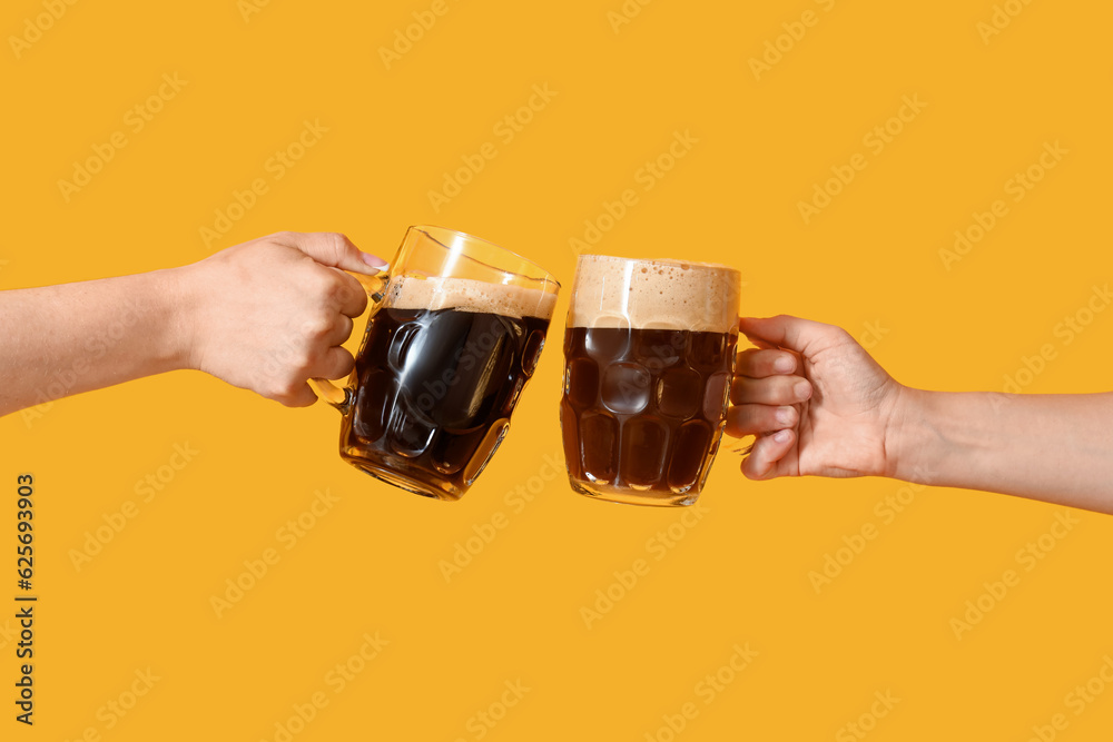 Female hands with mugs of cold beer clinking on yellow background