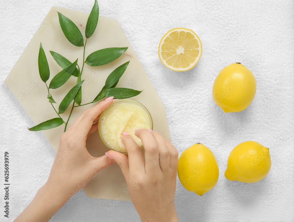 Female hands with bowl of lemon body scrub and towel on white tile background